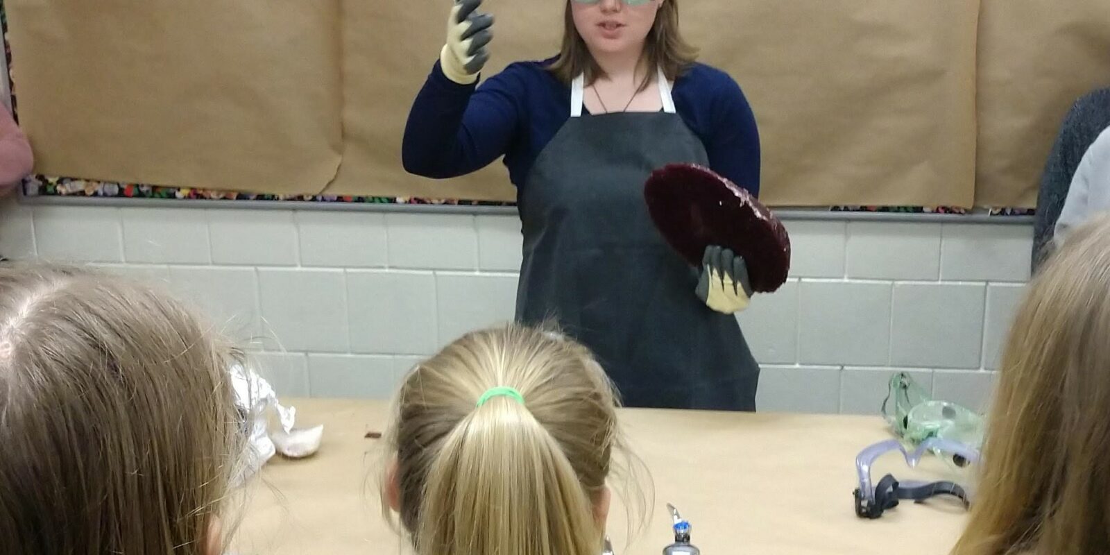 Volunteer, Erin Crowley, stands at the front of a classroom wearing safety goggles and gloves and holds a large piece of "obsidian" rock candy as she prepares to demonstrate to students how to flintknap.