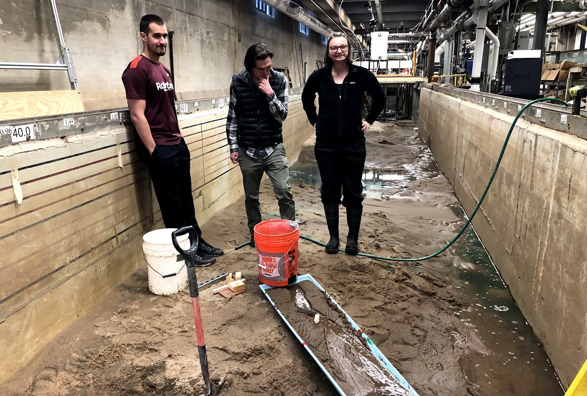 Three researchers stand in a cement channel (flume) and stand atop wet sediment in the research laboratory.