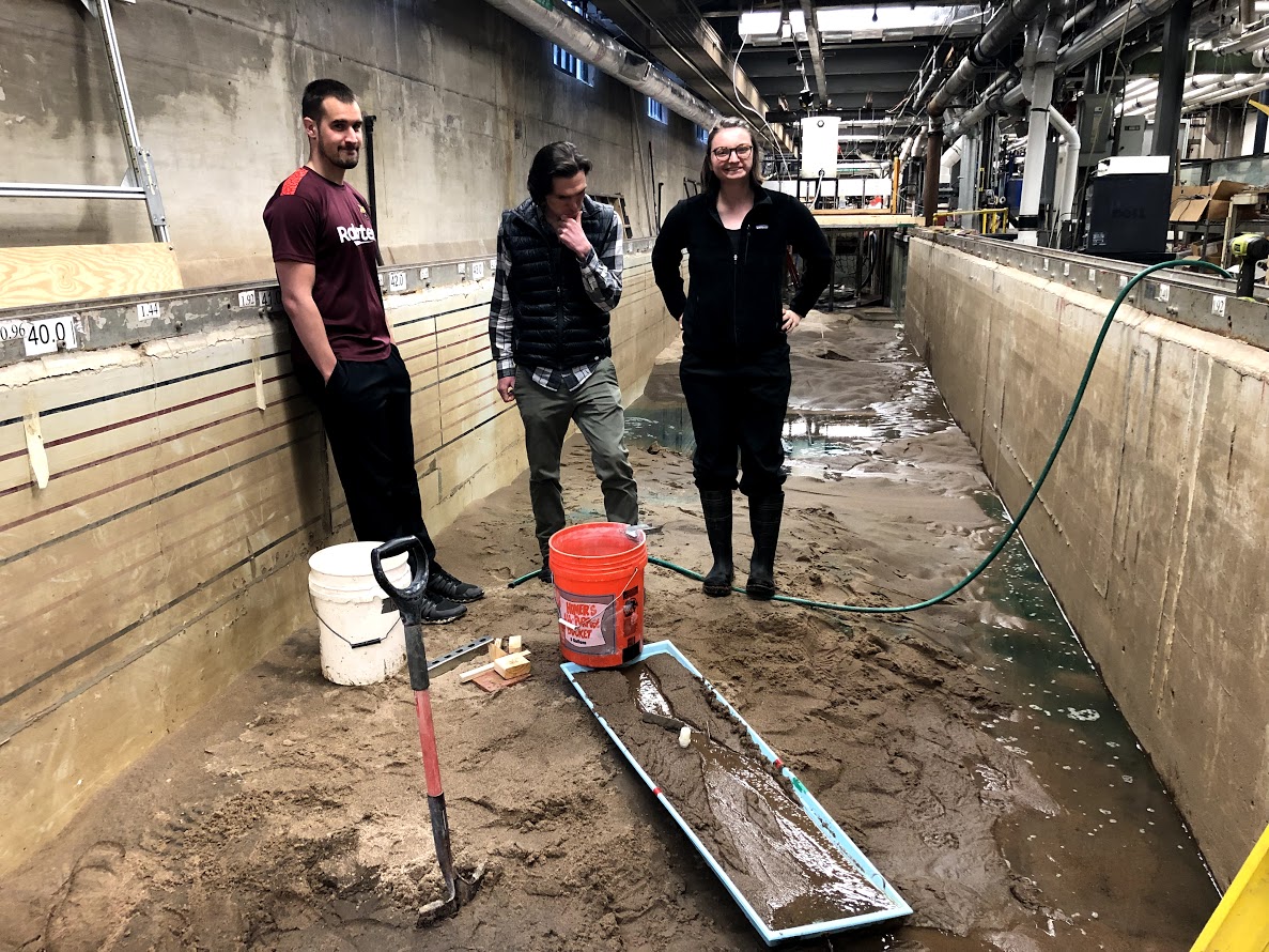Three researchers stand in a cement channel (flume) and stand atop wet sediment in the research laboratory.