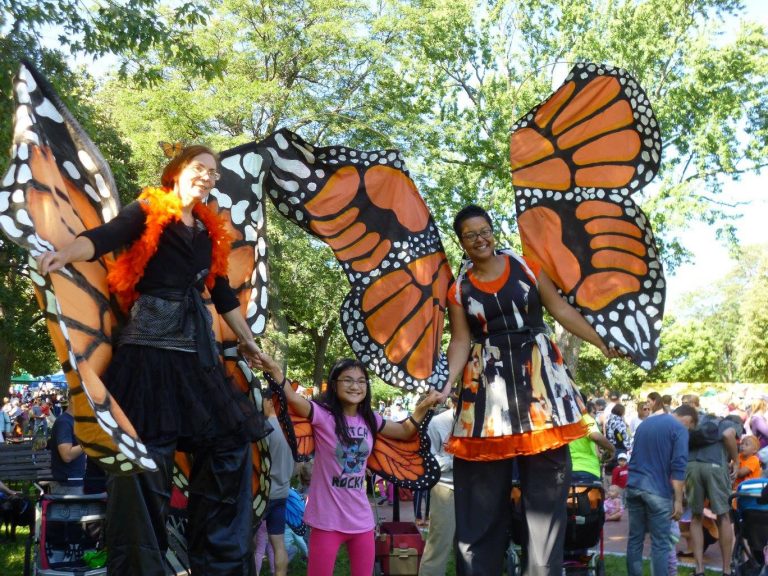 Photo showing two people standing high on stilts, dressed as monarch butterflies with large orange, black, and white wings, and an excited child, also wearing wings, standing between them.